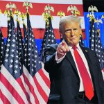 U.S. president Donald Trump stands, pointing directly at the camera with his right index finger, in front of a row of U.S. flags.