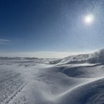 Snow is piled along the side of a road and the sun is low in the clear blue sky on a cold day in February in Saskatchewan.