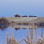 A large red tractor in the distance is pulling an air seeder and an anhydrous ammonia tank on wheels with a slough in the foreground.