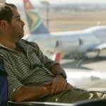 A weary traveller sits in an airport with planes on the tarmac visible through the windows in the background.