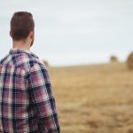 A young male farmer stares at large round bales in the distance.