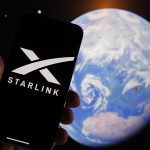 A hand holds a smart phone with the Starlink company logo on its screen while a photo of the blue and white earth from space is visible in the background.