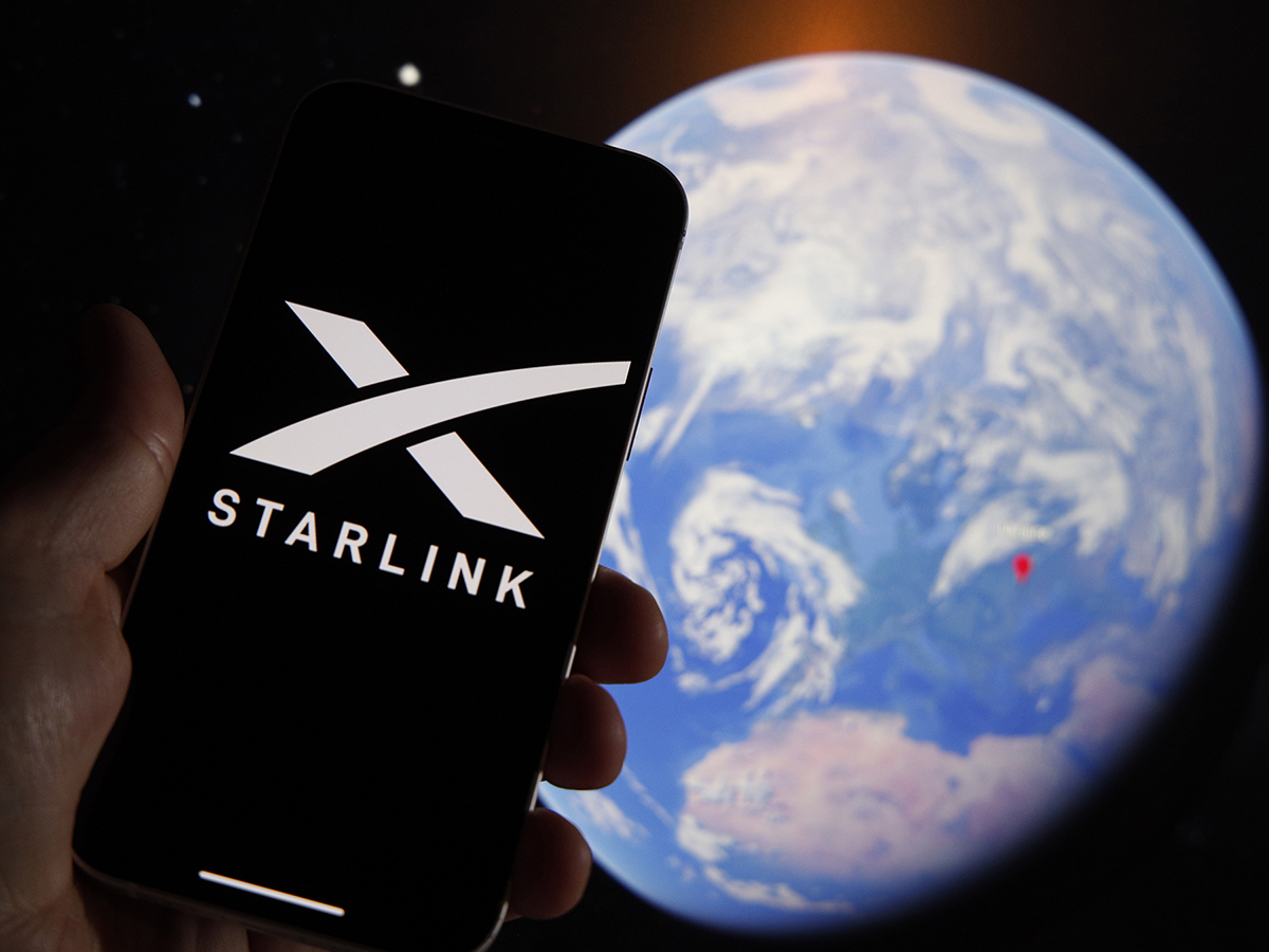A hand holds a smart phone with the Starlink company logo on its screen while a photo of the blue and white earth from space is visible in the background.