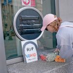 A person leaves a small boquet of flowers next to a sign drawn like a gravestone that says, RIP USAID - 1961 - 2025"