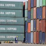 Multi-coloured shipping containers sit stacked 5-high on a dock, dawrfing three people walking on the pavement in front of them.