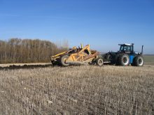 A large blue tractor pulls a soil scraper through an unplanted field.