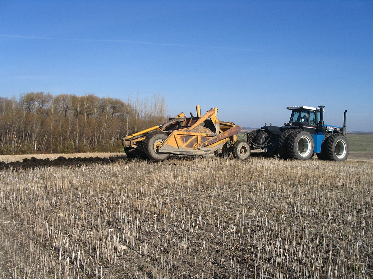 A large blue tractor pulls a soil scraper through an unplanted field.