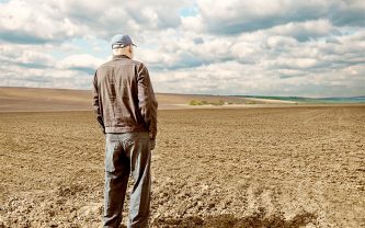 An elderly farmer stands in an unplanted field looking into the distance.