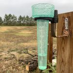 Water sits in the bottom of a rain gauge attached to a fence on a cloudy and rainy day.