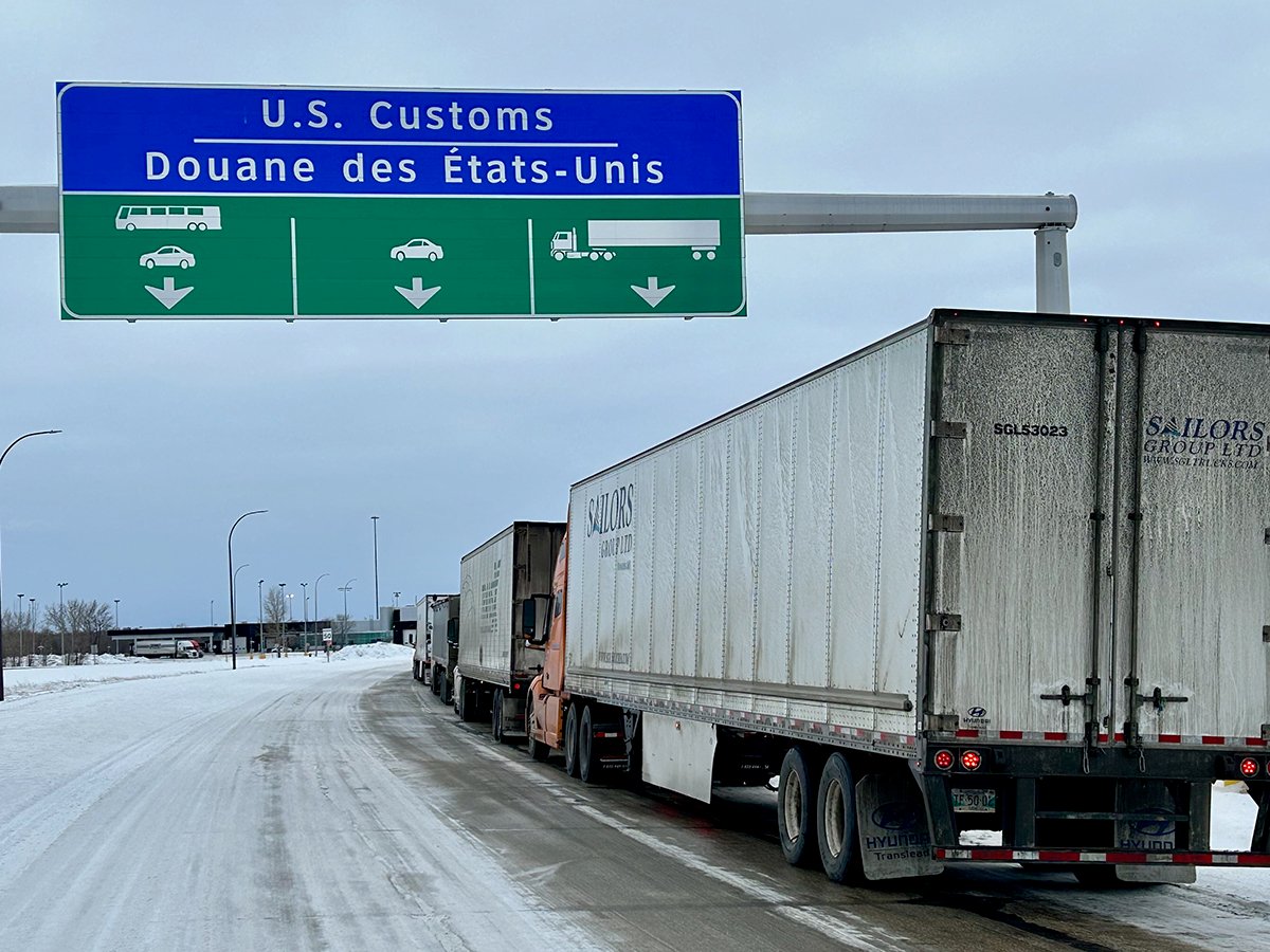 Semi trucks lined up on a highway in winter beneath a sign that reads, "U.S. Customs."