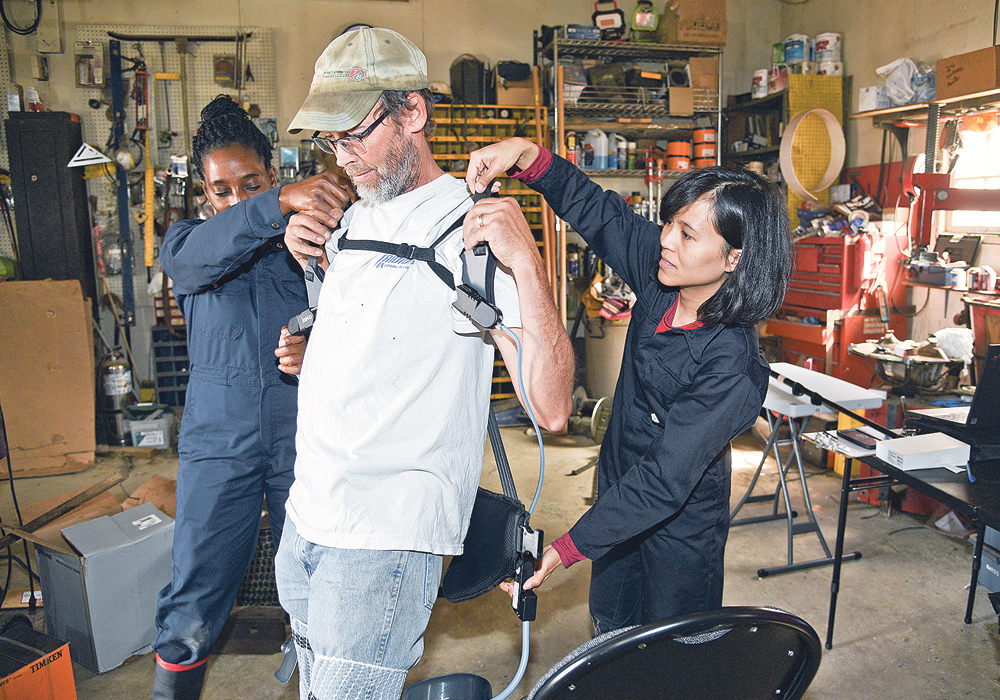 Abisola Omoniyi and Ornwipa Thamsuwan from the Canadian Centre for Health and Safety in Agriculture at the University of Saskatchewan hook an exoskeleton to Ron Swan of Glenside, Sask., who volunteered to test the unit on the farm.  |  William DeKay photo