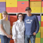 Krystal Braam, left, Freda Molenkamp-Oudman and Howard Oudman stand in front of colourful supers in the cold storage area at the farm’s honey house. Oudman is mentoring Braam in beekeeping. | Maria Johnson photo