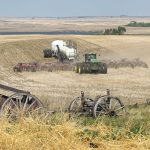 Photo looking down from the top of a hill with a modern air seeder rig approaching in the distance and some discarded old wooden equipment with wagon wheels in the foreground. with
