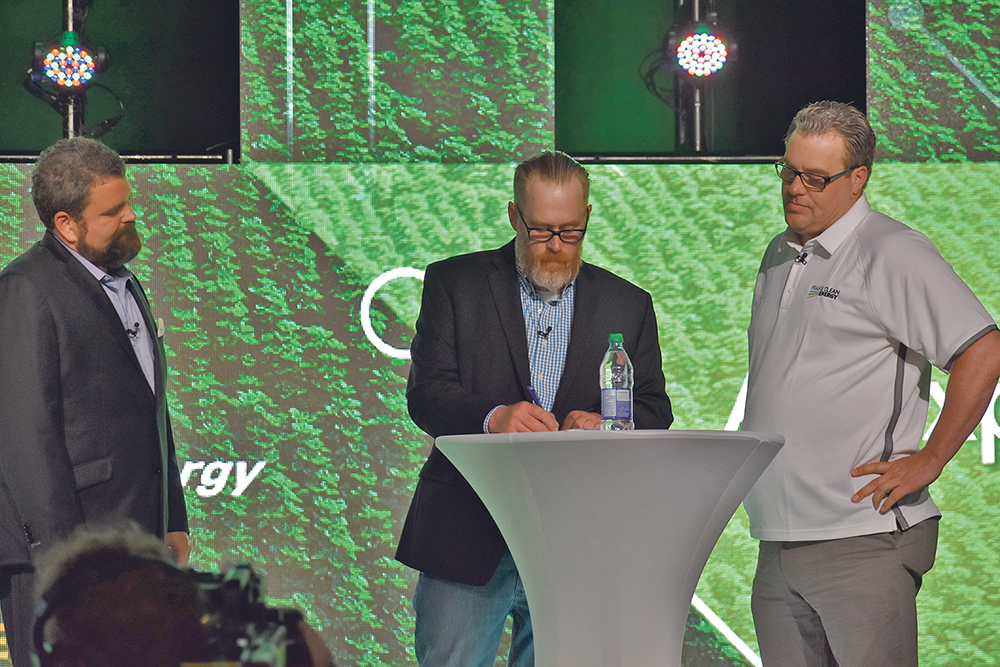 Mark Cooper, left, president of Prairie Clean Energy, and Trevor Thomas, right, the company’s chief relations officer, watch Thomas Mackenzie sign the first flax straw contract for the company’s new flax straw biomass plant.  |  Braedyn Wozniak photo