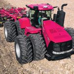 Drone shot from above and in front of a large, 8-tired red tractor towing a cultivator.