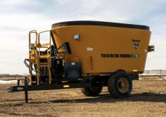 A towed, yellow vertical feed mixer sits parked, uattached to a tractor.