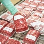 Close-up of a female shopper's hand lifting up a package of ground beef from a grocery store's meat display.