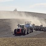 A large tractor with 8 tires pulls an air seeder down a hill, while a far truck carrying seed is parked atop the hill in the background.