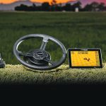 A promotional photo of a small control box, a steering wheel, a small computer screen, and a rooftop receiver all sitting on top of a hay bale.