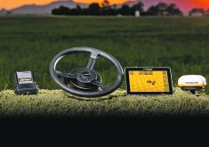 A promotional photo of a small control box, a steering wheel, a small computer screen, and a rooftop receiver all sitting on top of a hay bale.