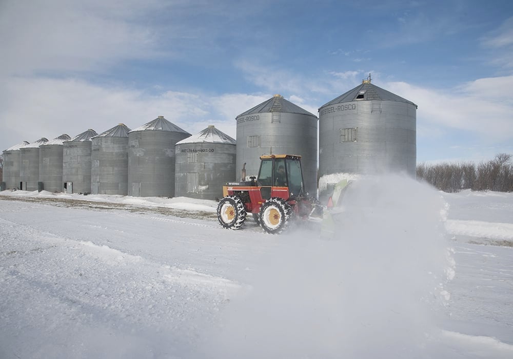 A tractor with a snowblower mounted on it clears snow from in front of steel grain bins.