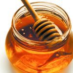 A honey stick rests on the surface and edge of a jar of liquid honey.