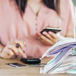A woman sits at a desk looking at her phone and punching numbers into a calculator. Credit or debit cards are on the table in front of her, as well as a stack of bills or cancelled cheques stuck on a spike, and an alarm clock.