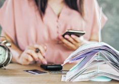 A woman sits at a desk looking at her phone and punching numbers into a calculator. Credit or debit cards are on the table in front of her, as well as a stack of bills or cancelled cheques stuck on a spike, and an alarm clock.