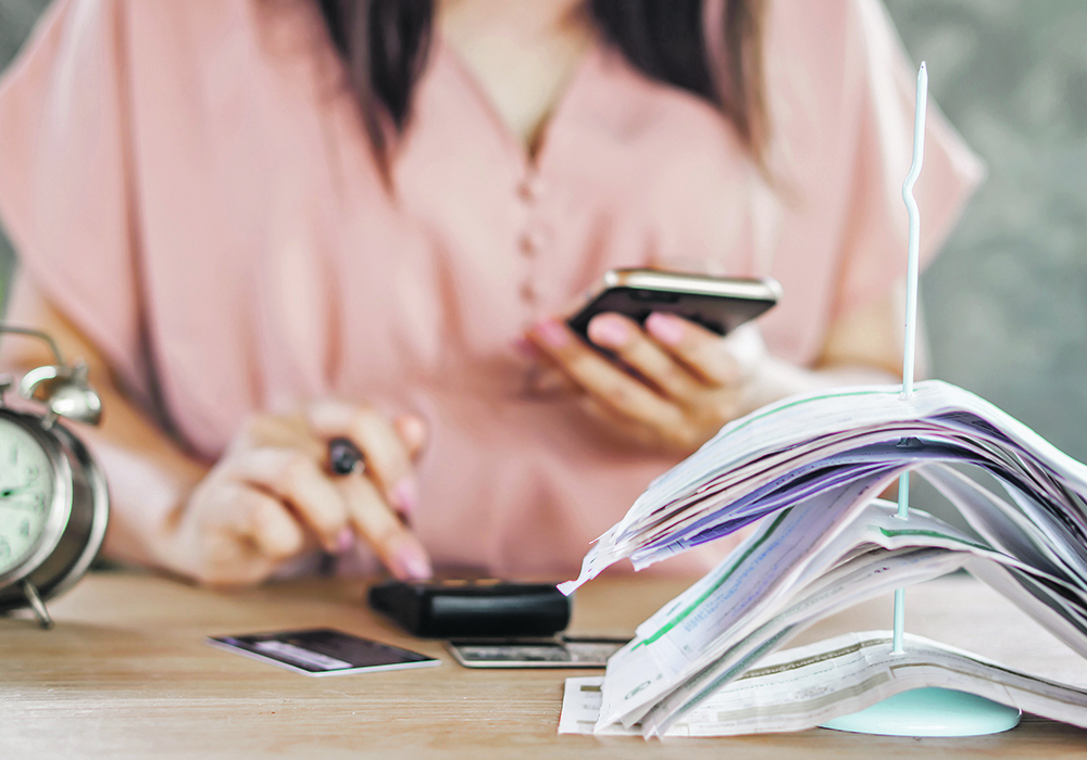 A woman sits at a desk looking at her phone and punching numbers into a calculator. Credit or debit cards are on the table in front of her, as well as a stack of bills or cancelled cheques stuck on a spike, and an alarm clock.