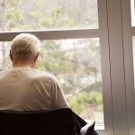 An elderly man sits in a chair looking out a window.