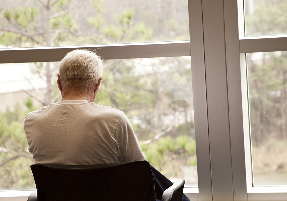 An elderly man sits in a chair looking out a window.