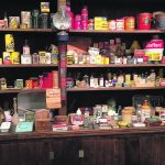 Photo of old fashioned product shelving from a general story chock full of products, with two lanterns and powder-filled fire extinguishers hung beneath.