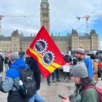 Public Service Alliance of Canada workers picket on Parliament Hill in Ottawa. Two picketers carry a PSAC flag in the foreground, with the Centre Block in the background.