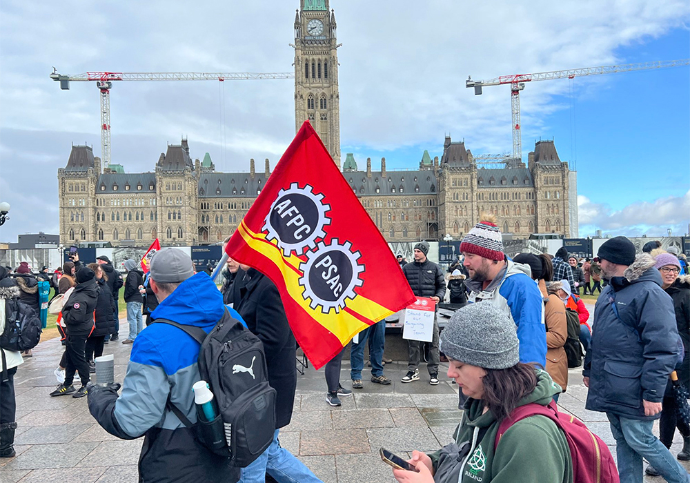 Public Service Alliance of Canada workers picket on Parliament Hill in Ottawa. Two picketers carry a PSAC flag in the foreground, with the Centre Block in the background.