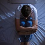 An overhead shot of a young man sitting on his bed in the dark hugging his knees to his chest.