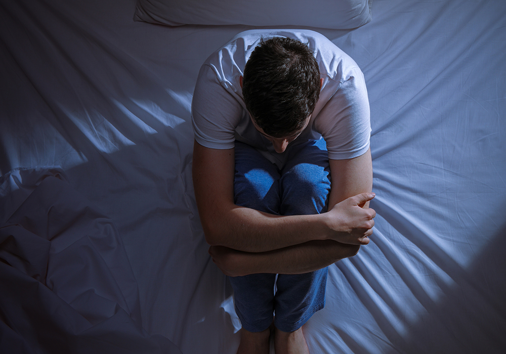 An overhead shot of a young man sitting on his bed in the dark hugging his knees to his chest.
