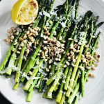 An overhead shot of a plate of asparagus sprinkled with pine nuts and parmesan and a lemon wedge on the side of the plate.
