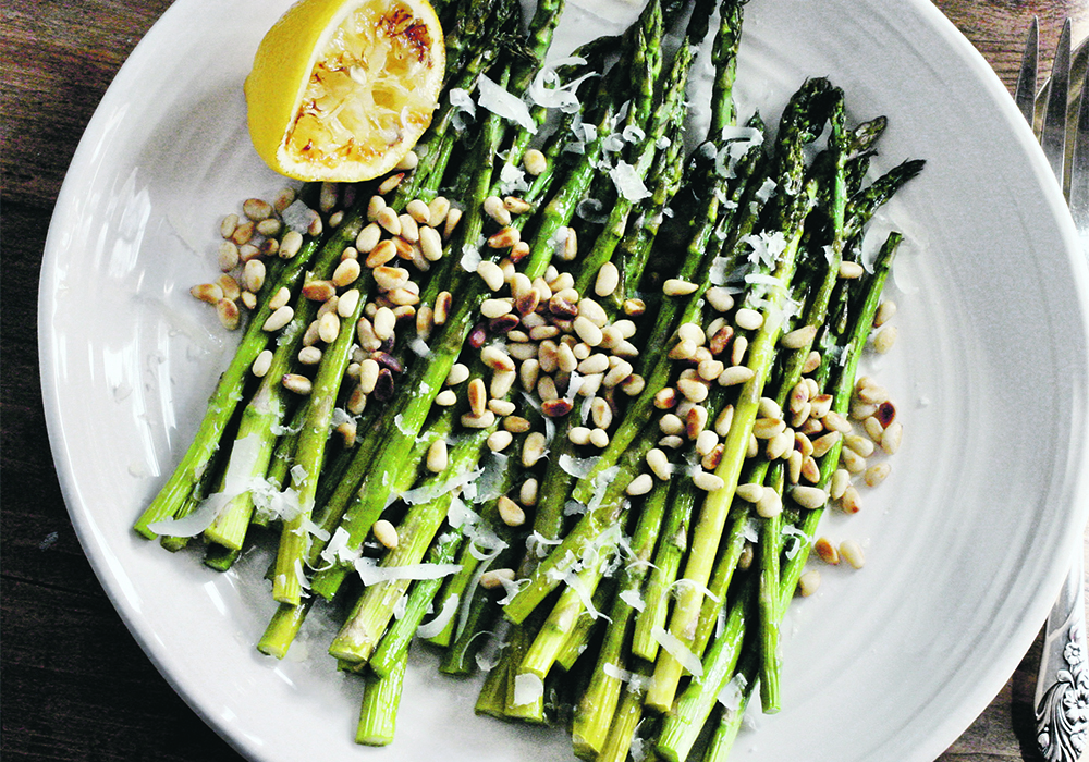 An overhead shot of a plate of asparagus sprinkled with pine nuts and parmesan and a lemon wedge on the side of the plate.