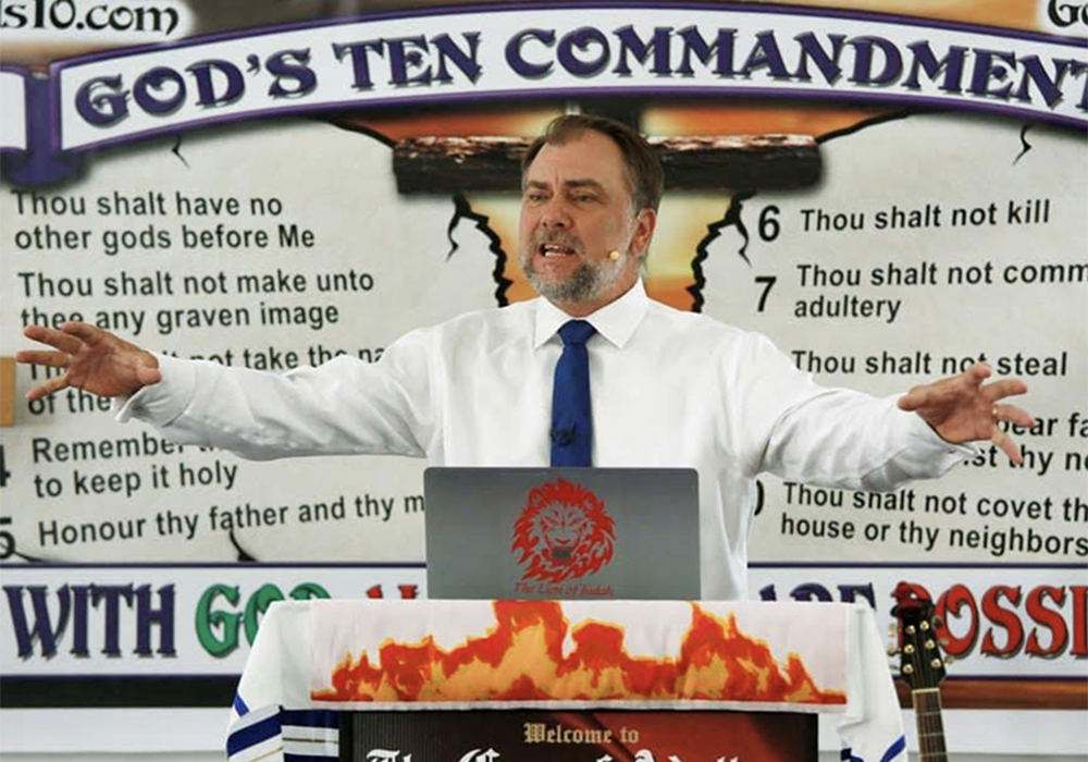 Artur Pawlowski preaches from a lectern with the "God's Ten Commandments" on the wall behind him.