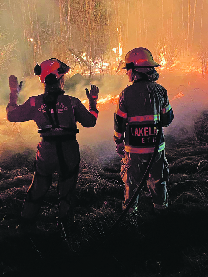 An experienced firefighter explains something to a student firefighter from Lakeland College. Burning trees and grass are in the background, just a few meters away.