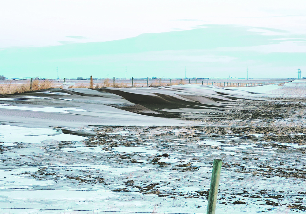 Soil is blown into piles near a fence line, there is some snow on top.