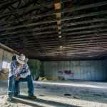 A young man dressed in jeans and a cowboy hat sits alone on a stool on the concrete floor of what appears to be a barn of some type. He's holding his hand up to his face, and the brim of his hat prevents us from seeing his face.