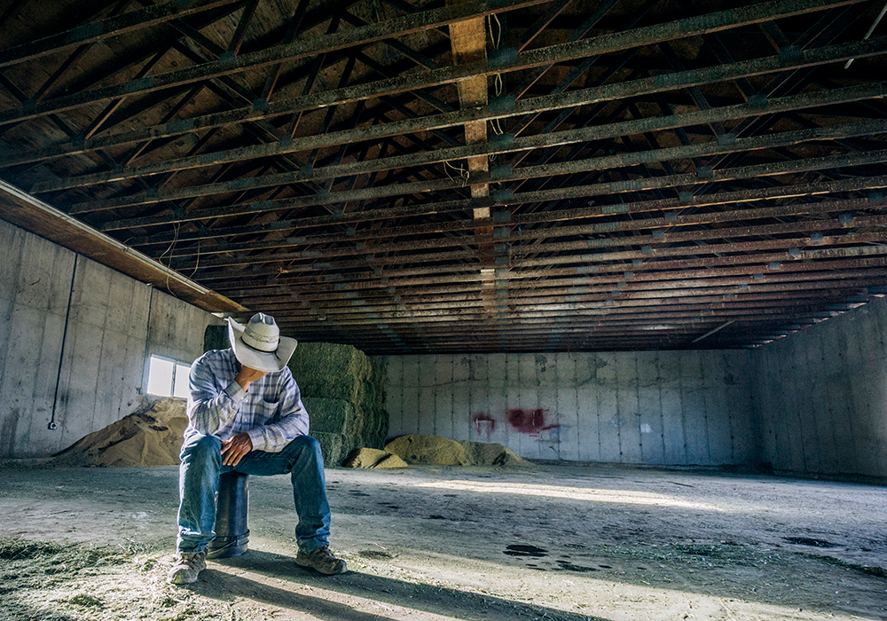 A young man dressed in jeans and a cowboy hat sits alone on a stool on the concrete floor of what appears to be a barn of some type. He's holding his hand up to his face, and the brim of his hat prevents us from seeing his face.