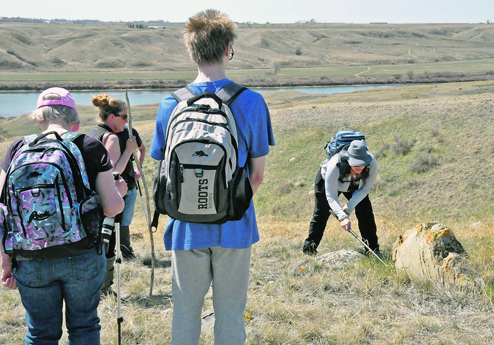 A woman uses a snake hook to carefully turn over a rock that may have snakes beneath it while school kids look on.