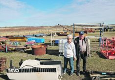 An older man and his wife stand outside among some of the items listed on their online auction.