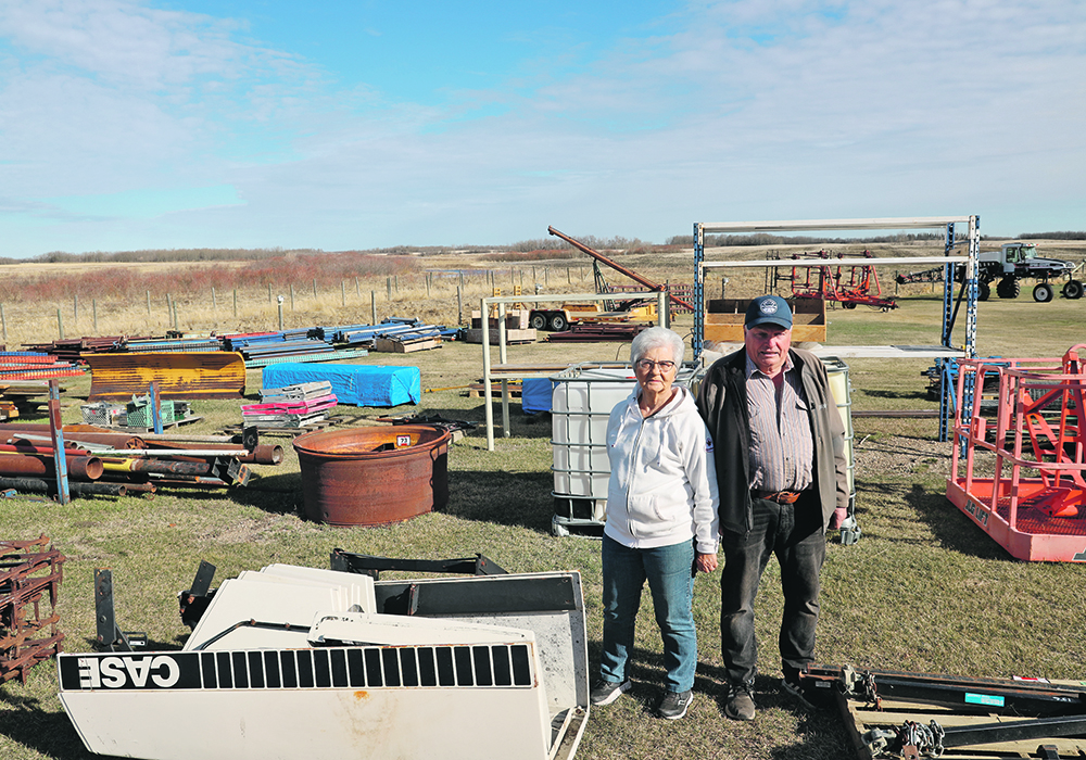 An older man and his wife stand outside among some of the items listed on their online auction.
