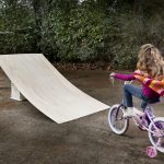 A young girl on a bike with training wheels rides toward a ramp with a little boy seated on the ground in front of the ramp.