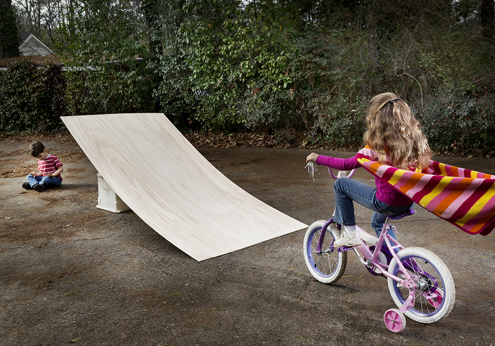 A young girl on a bike with training wheels rides toward a ramp with a little boy seated on the ground in front of the ramp.