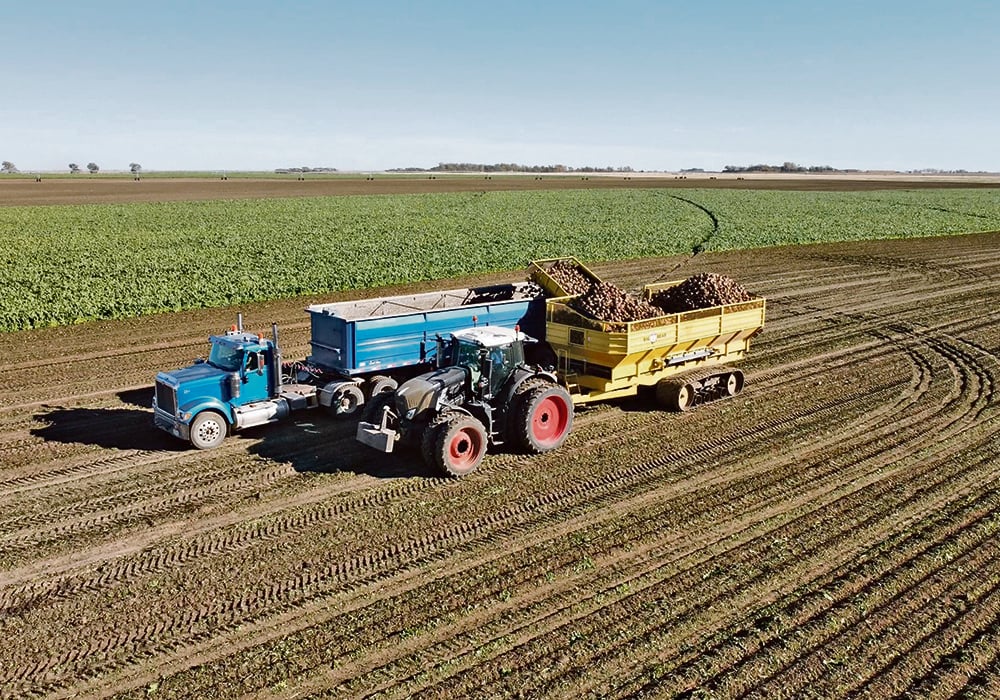 An overhead drone photo of a tractor pulling a hopper filled with beets transferring them to a semi trailer for transport from the field.