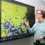 A researcher stands in front of a big screen tv displaying satellite imagery of farmland.